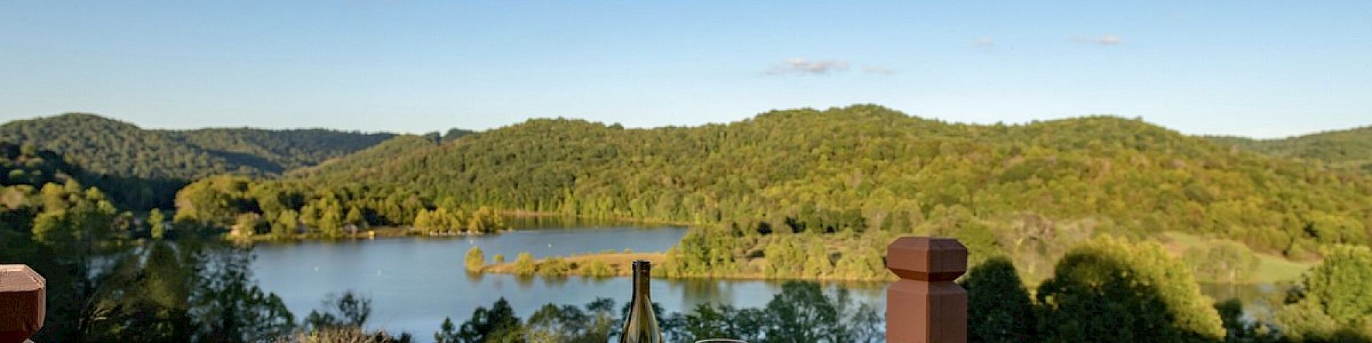 A wooden table with wine, glasses, and plates of food on an outdoor deck overlooking a scenic lake and forested hills under a clear blue sky.