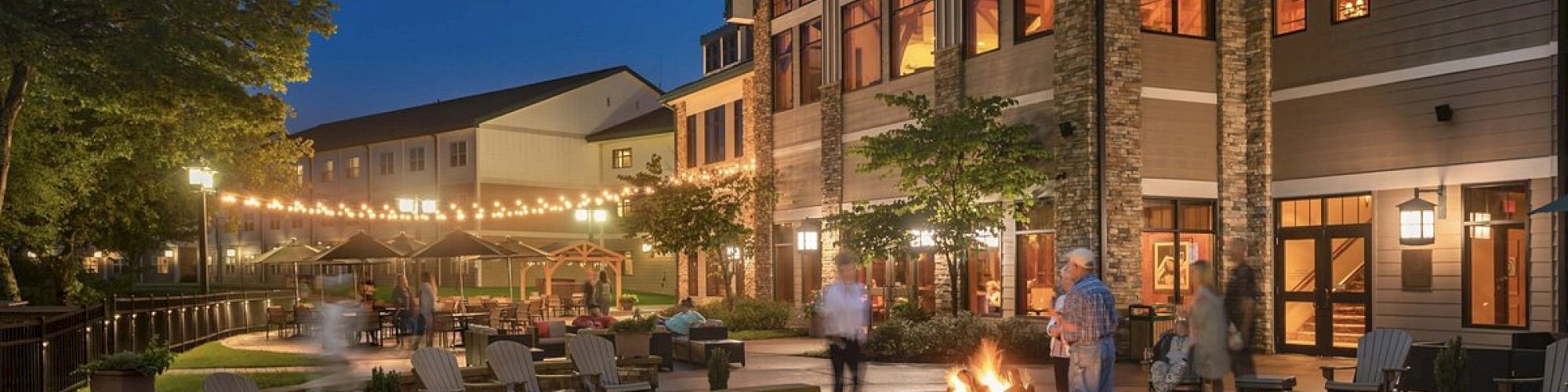 An evening scene of a cozy, spacious patio at a resort with a fire pit, Adirondack chairs, string lights, and several people enjoying the ambiance.