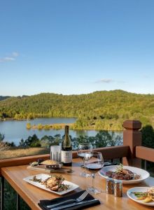 A table set with meals and wine overlooks a serene landscape with a lake and forested hills under a clear, blue sky.