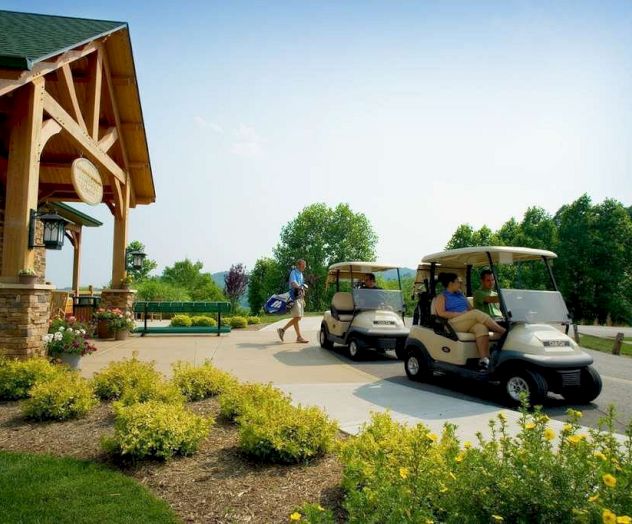 A person walks with golf clubs towards a clubhouse, while two people sit in golf carts near landscaped greenery on a sunny day.