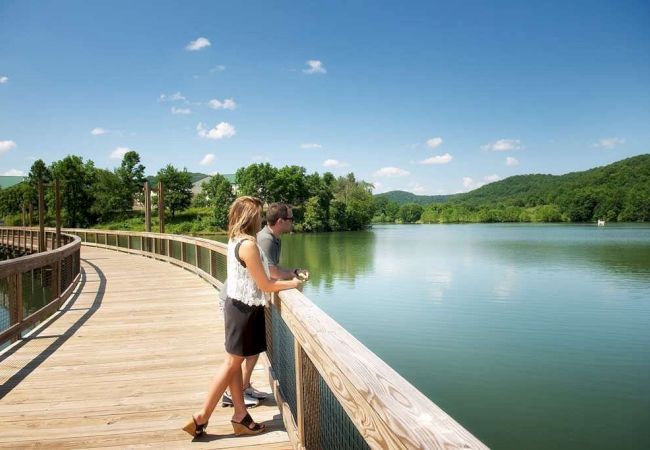 A couple stands on a wooden boardwalk overlooking a serene lake with green hills in the background, enjoying a sunny day in nature.