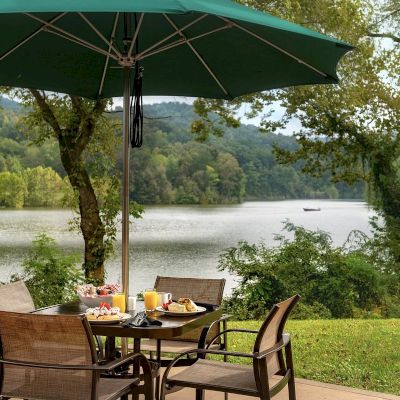 An outdoor dining setup with a table and chairs under a green umbrella, overlooking a scenic lake and lush greenery, with plates of food.