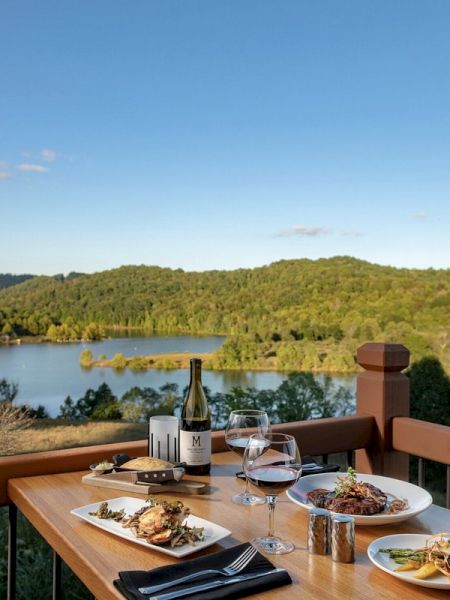 A table set with plates of food, wine, and glasses on a balcony with a scenic view of a lake and wooded hills in the background.