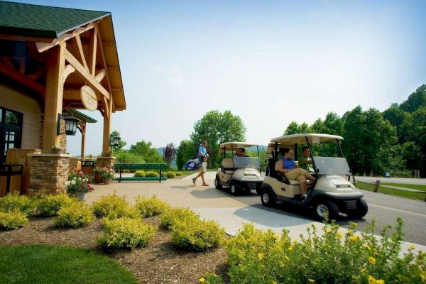 A person walks toward a building carrying golf clubs, while two people sit in golf carts nearby on a sunny day surrounded by greenery.