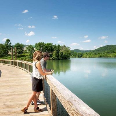 A couple stands on a wooden boardwalk overlooking a serene lake surrounded by lush green hills and trees under a clear blue sky.