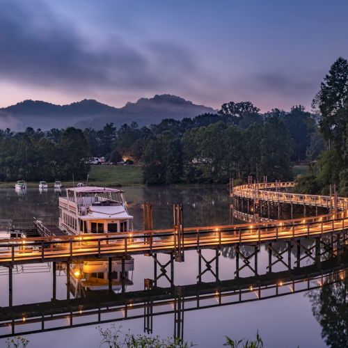 A scenic view of a wooden boardwalk over a calm river, winding through trees at dusk with mountains in the background and a boat docked along it.