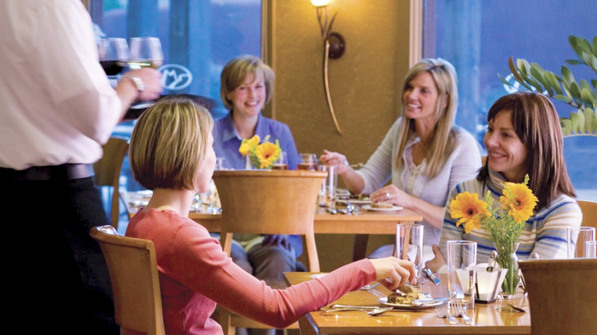 A waiter serves a group of four people seated at a table in a restaurant, with sunflowers as centerpiece and outdoor scenery visible through the window.