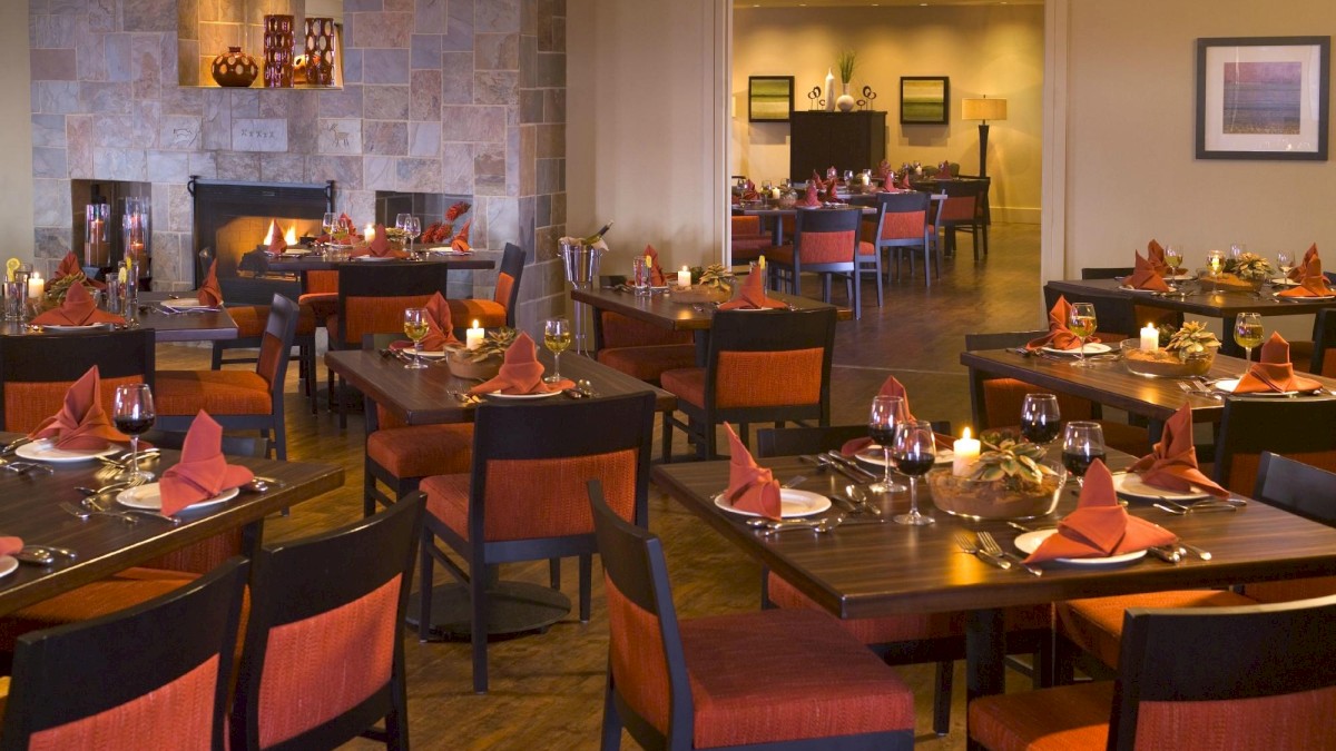 A cozy restaurant with neatly arranged tables and red napkins, dim lighting, and a welcoming atmosphere. A fire glows in the background fireplace.