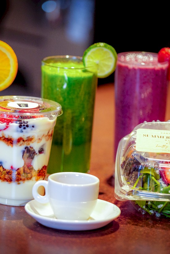 The image shows three colorful smoothies, a yogurt parfait, a salad in a container, and a small white cup on a brown countertop.