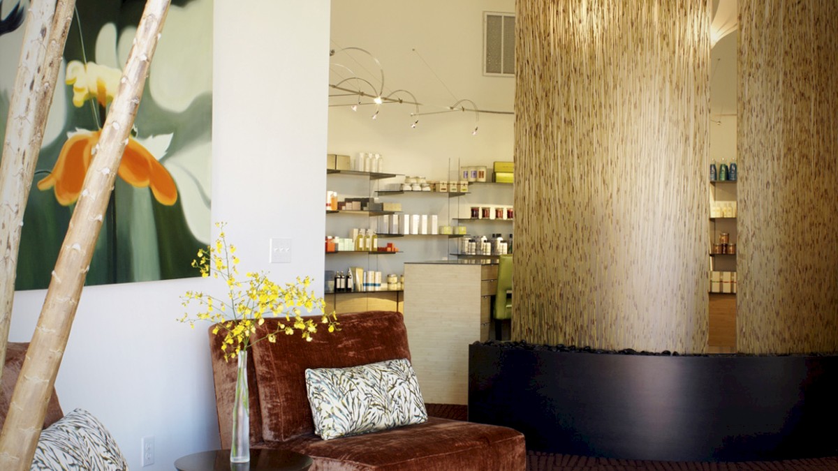 A modern interior with plush chairs, a decorative vase, a wall painting, a cylindrical room divider, and shelves of various products in the background.