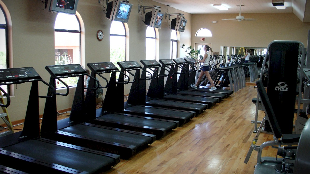 A gym with several treadmills lined up facing a wall-mounted TV screen and mirrors, with one person using a treadmill.