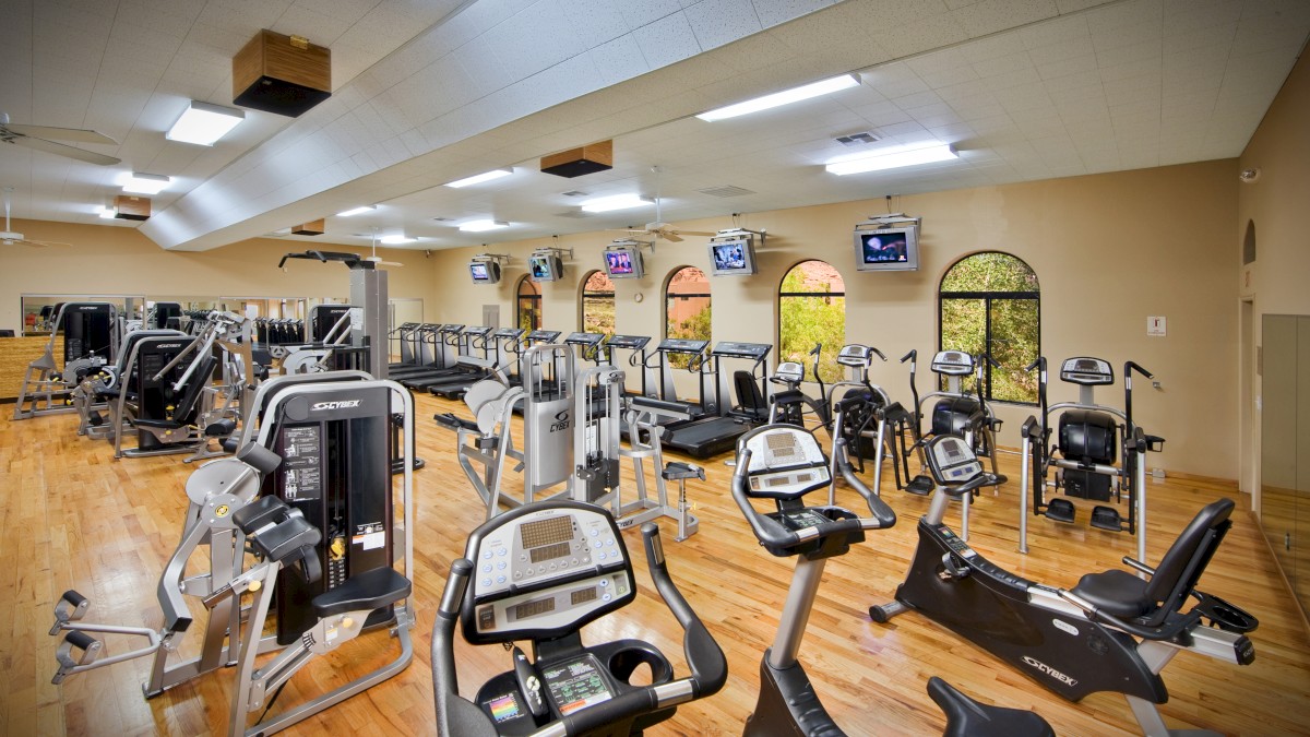 This image features a well-equipped gym with various exercise machines, including treadmills, stationary bikes, and weight machines, on a wooden floor.