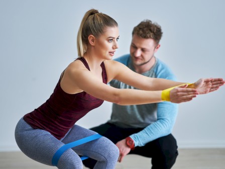 A woman is performing a squat exercise with resistance bands while a man, possibly a trainer, supervises her form, in an indoor setting.