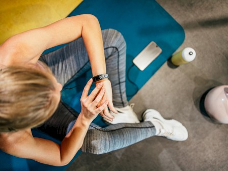 A person in workout attire is sitting on a blue mat, checking their smartwatch. Nearby are a smartphone, water bottle, and kettlebell.