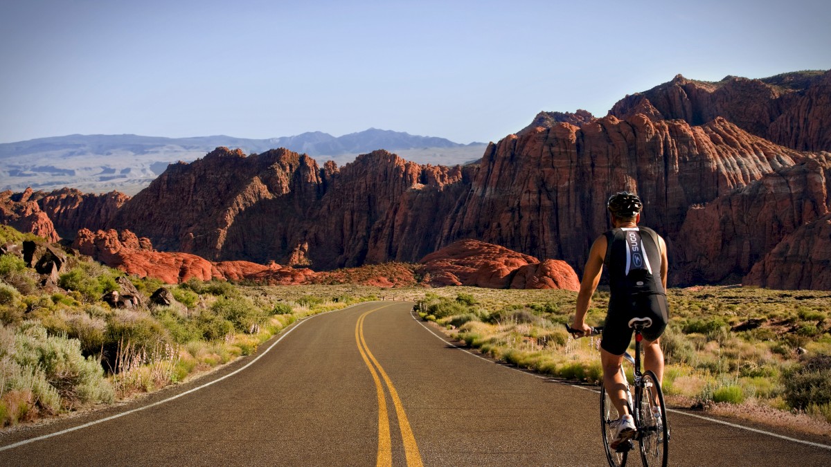 A cyclist rides on an empty road towards rugged, red rock mountains under a clear sky, surrounded by desert vegetation.