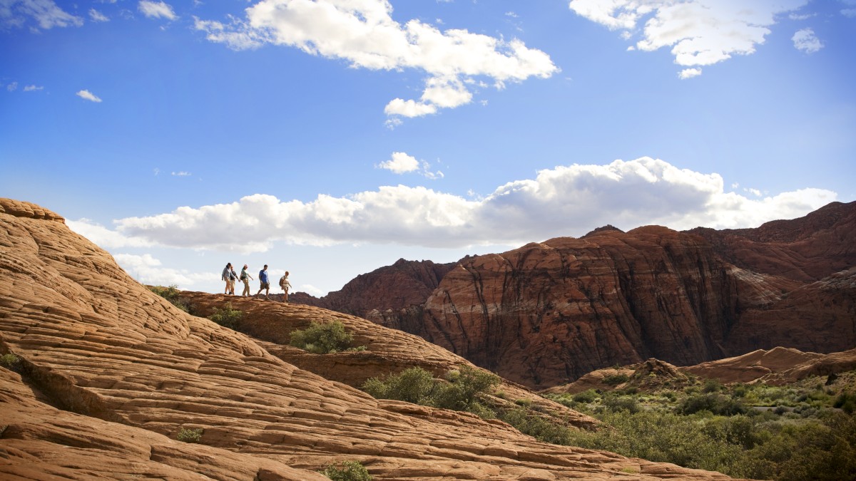 A group of people is hiking on a rocky terrain with mountains in the background, under a bright blue sky with scattered clouds.