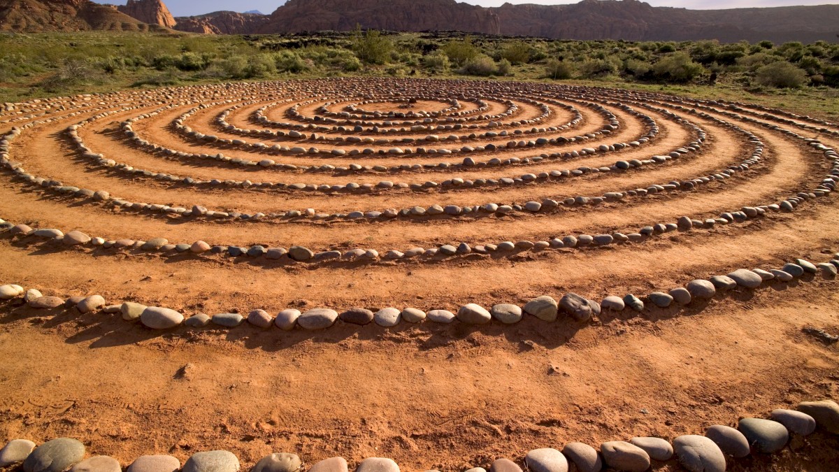 The image shows a spiral labyrinth made of circular paths outlined with stones in a desert landscape, with mountains and sparse vegetation in the background.