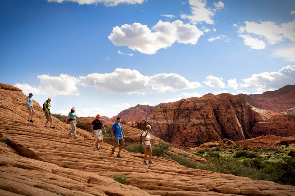 A group of people are hiking on a red rocky terrain under a blue sky with scattered clouds and a backdrop of mountains.