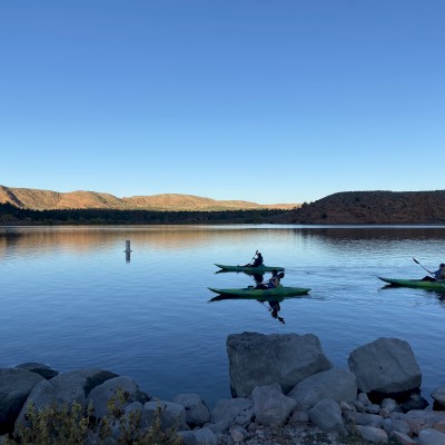 Three kayakers paddle on a calm lake with hilly terrain and clear blue skies in the background.