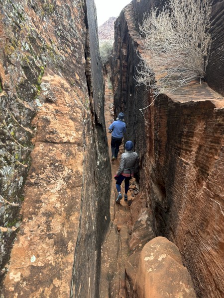 Two people are walking through a narrow, rocky canyon. The walls are steep and covered in different shades of brown and orange rock.