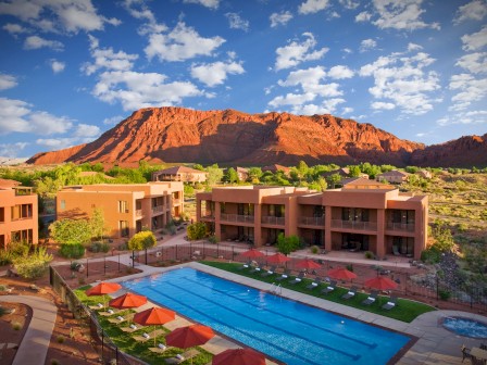 A resort features an outdoor pool with red umbrellas, surrounded by desert vegetation and overlooking a dramatic red rock mountain under a blue sky.