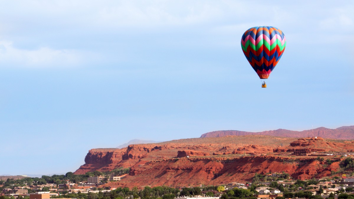 A colorful hot air balloon floats above a desert landscape with mesas and a town below it, under a clear sky.