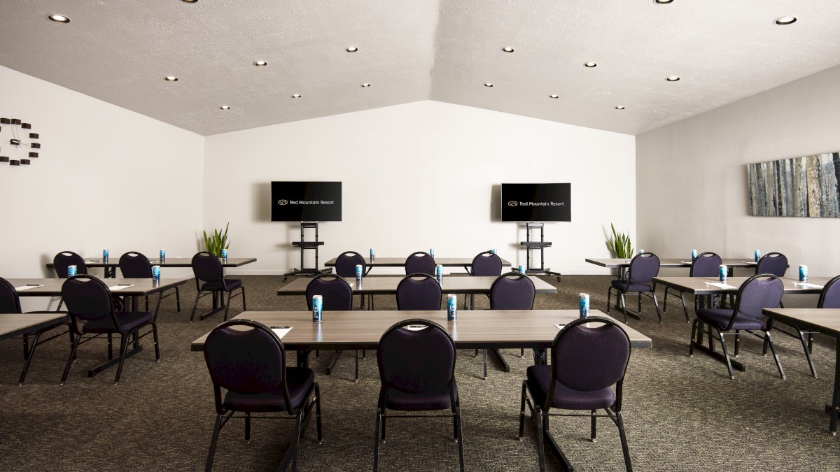 A conference room with multiple tables and chairs, two large screens at the front, plants, and beverage cans placed on the tables in the image.