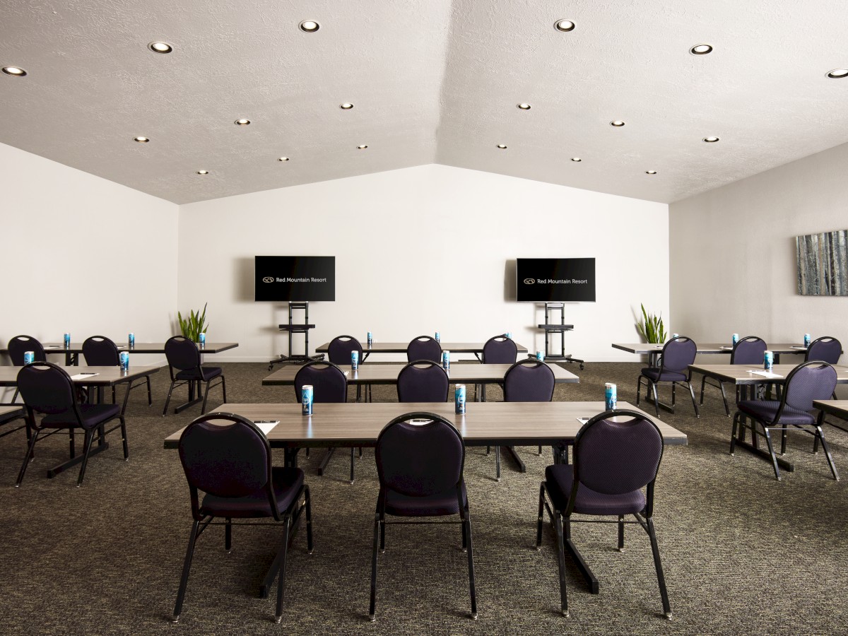 A conference room with multiple tables and chairs, two large screens at the front, plants, and beverage cans placed on the tables in the image.