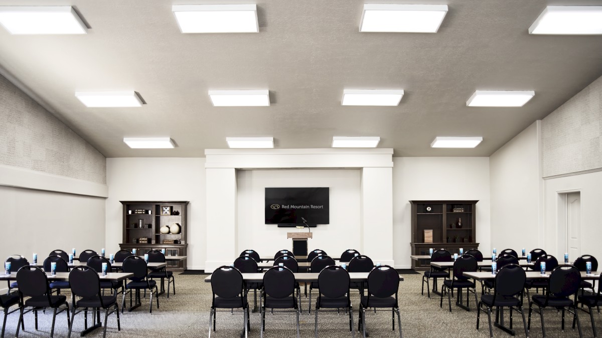 The image depicts a conference room with rows of tables and chairs arranged for a meeting or presentation, facing a podium and screen.