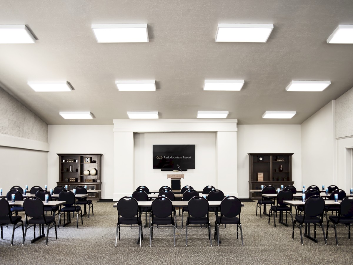 The image depicts a conference room with rows of tables and chairs arranged for a meeting or presentation, facing a podium and screen.