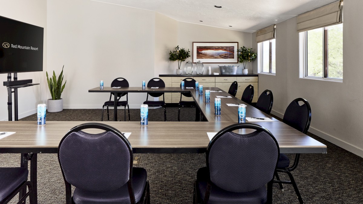 The image shows a conference room with tables arranged in a U-shape, chairs, water bottles, a screen, and windows with natural light.