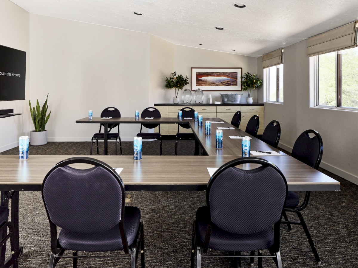 The image shows a conference room with tables arranged in a U-shape, chairs, water bottles, a screen, and windows with natural light.