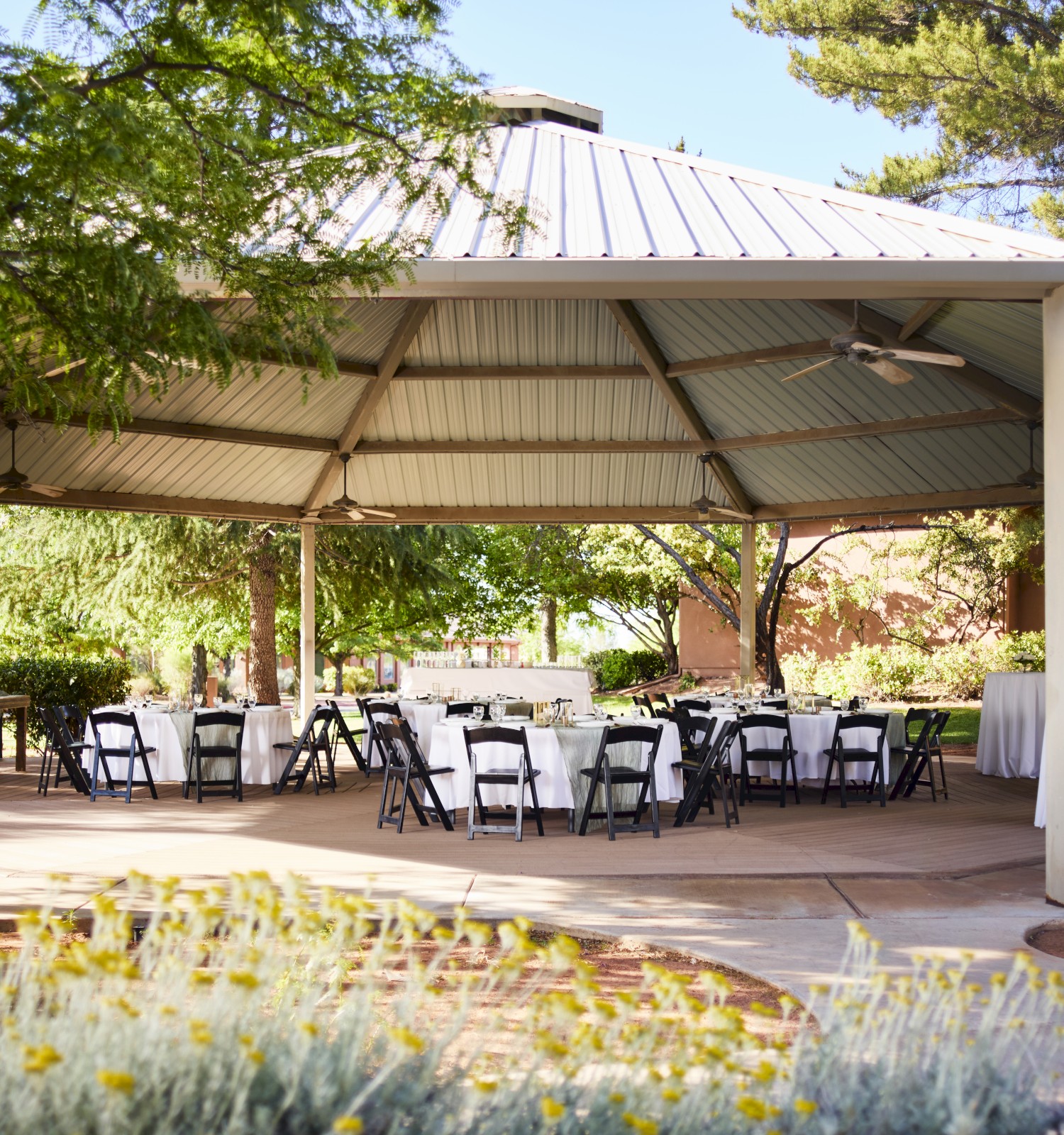 A gazebo with a metal roof is set up for an event with round tables and chairs, surrounded by greenery and flowers.