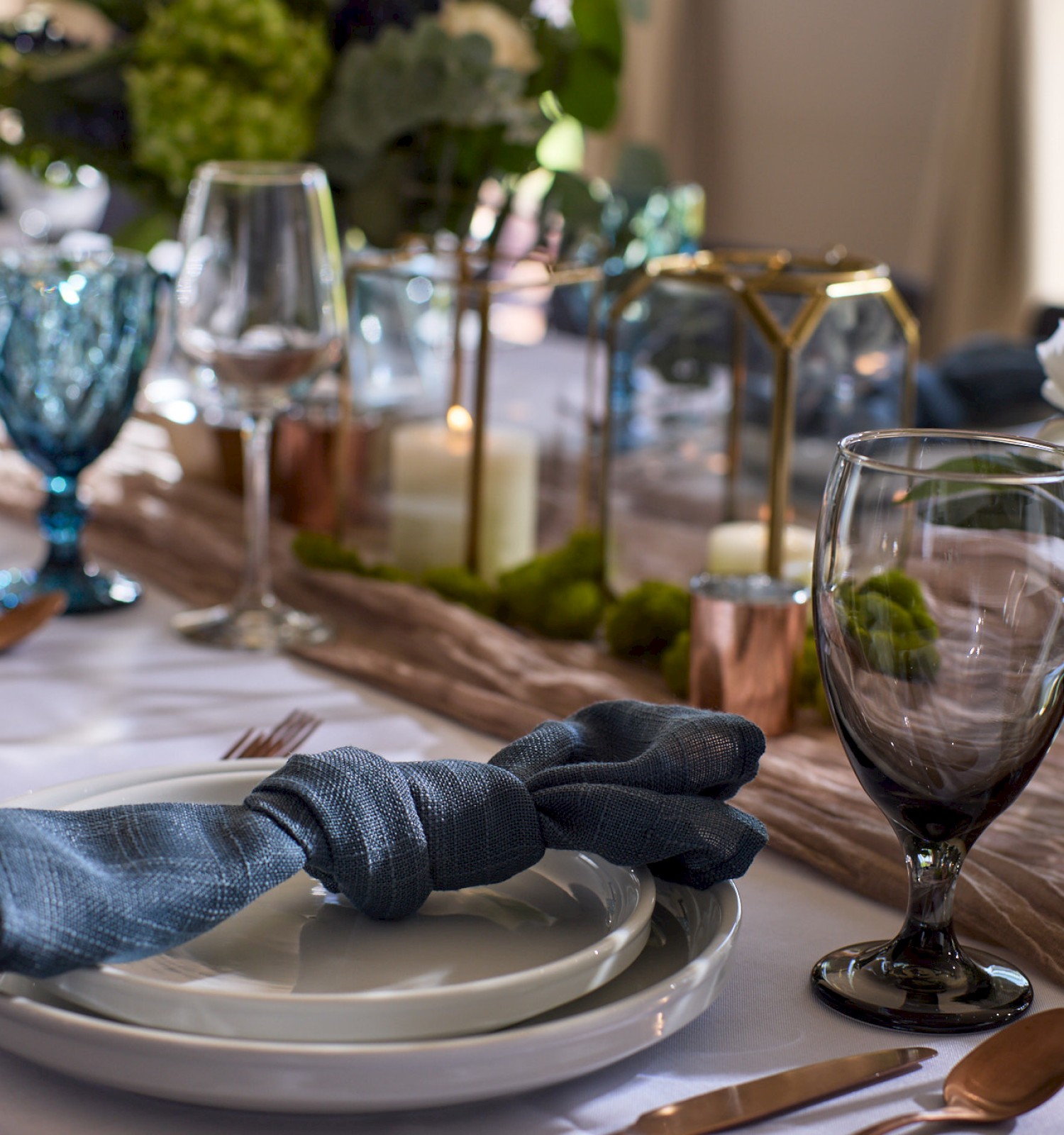 A beautifully set table with glassware, plates, blue napkins, and centerpieces featuring greenery and candles in glass and brass holders.