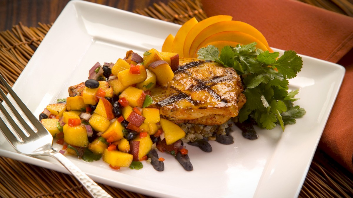 A plate of grilled chicken breast topped with a fruit salsa, accompanied by greens and garnished with orange slices, served with a fork.