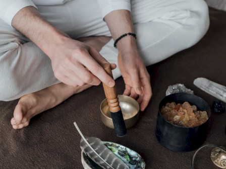 A person dressed in white sits crossed-legged, using a Tibetan singing bowl surrounded by spiritual and cleansing items like sage, crystals, and incense.