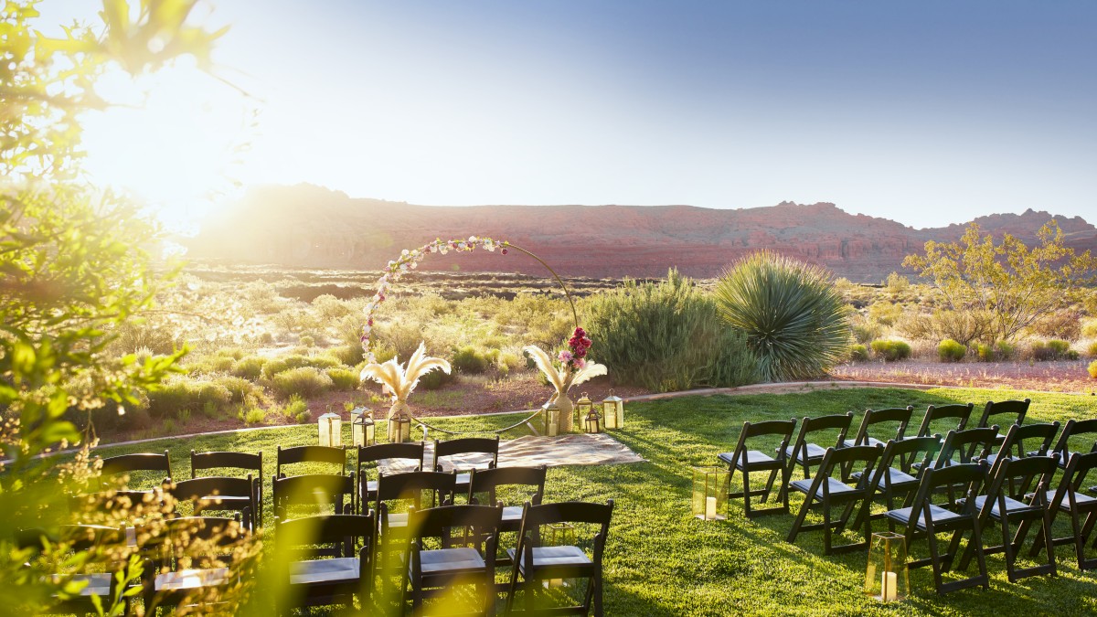 An outdoor event setup with rows of black chairs facing a decorated area, surrounded by greenery and a scenic landscape at sunset.