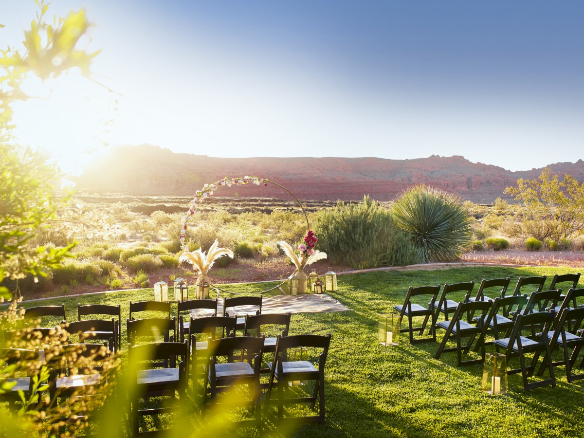 An outdoor event setup with rows of black chairs facing a decorated area, surrounded by greenery and a scenic landscape at sunset.