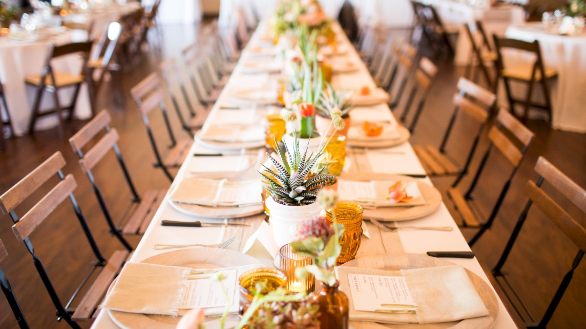 A long, elegantly set dining table with utensils, napkins, plates, and floral centerpieces in a well-lit room with wooden chairs around it.