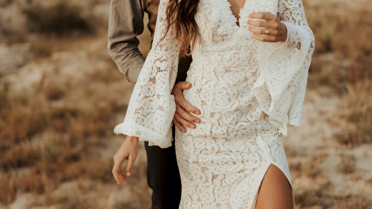 A couple is embracing, with the woman in a white lace dress and the man partially visible behind her, in an outdoor setting.