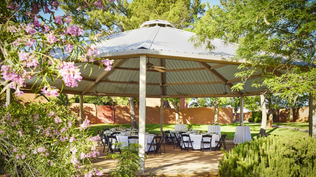 An outdoor pavilion with a metal roof is set up with tables and chairs, surrounded by trees and blooming flowers, in a park-like setting.