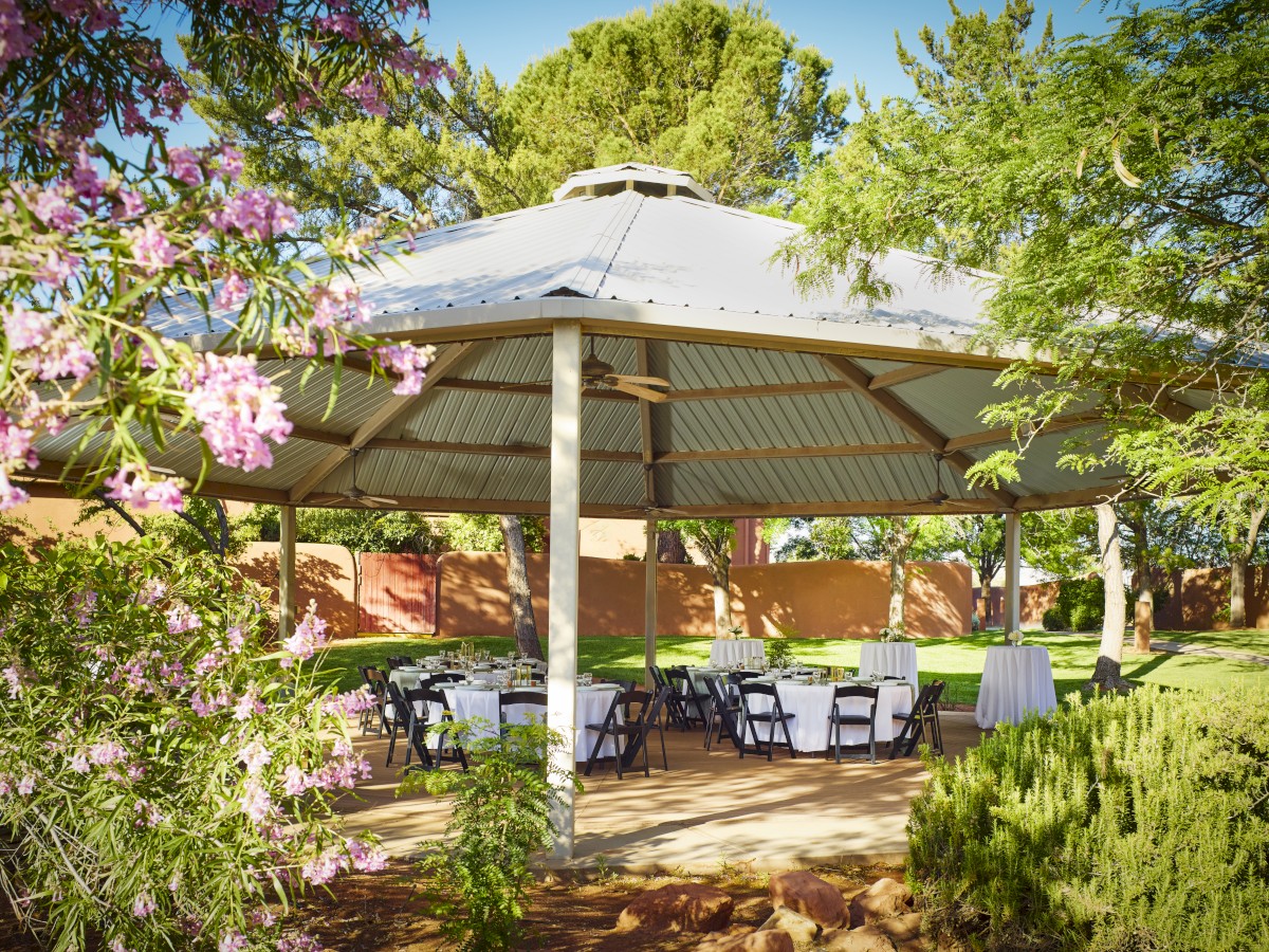 An outdoor pavilion with a metal roof is set up with tables and chairs, surrounded by trees and blooming flowers, in a park-like setting.