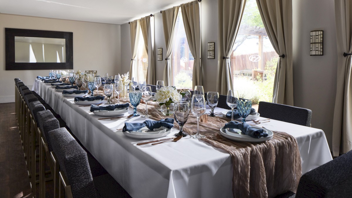 A long dining table set for a formal event with elegant tableware, glasses, and floral decorations next to large, curtained windows.