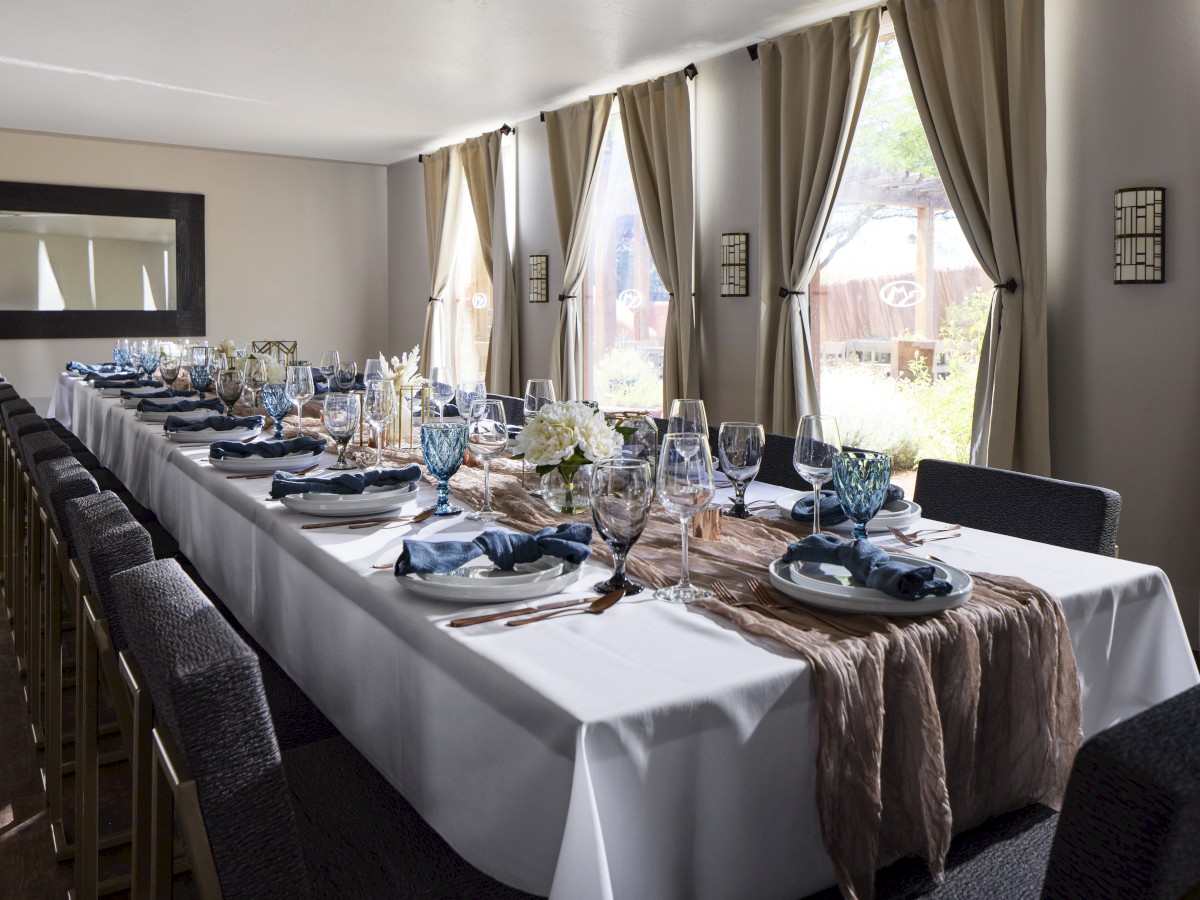 A long dining table set for a formal event with elegant tableware, glasses, and floral decorations next to large, curtained windows.