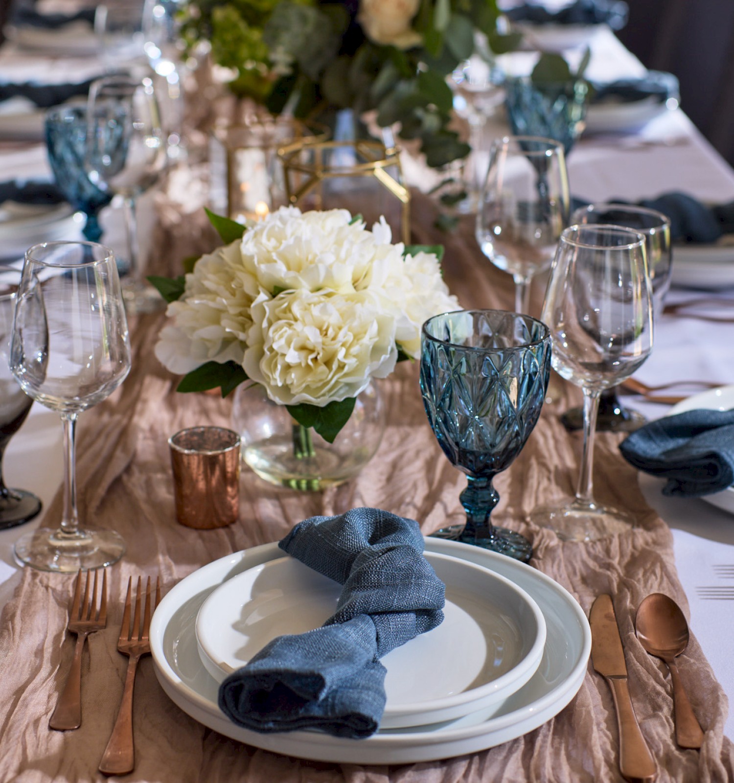 A beautifully set dining table featuring elegant blue glasses, white plates, neatly folded napkins, and a centerpiece of white flowers.