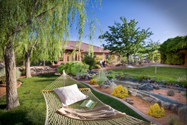 A peaceful garden with a hammock under a tree, near a stream, with a house in the background and a book on the hammock.