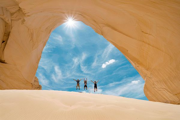 Three people are jumping in a sandy desert with a striking rock arch overhead and a bright sun shining through the opening.