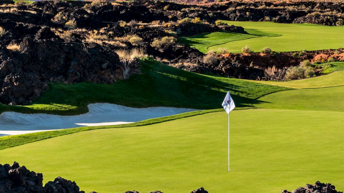 A lush golf course nestled among rugged rock formations with a sand bunker and a green flagstick, set against a backdrop of red mountains and blue sky.