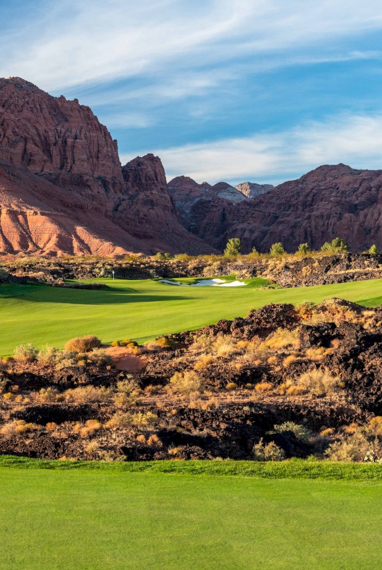 A lush green golf course is set against a backdrop of rugged red rock mountains under a bright, partly cloudy sky.