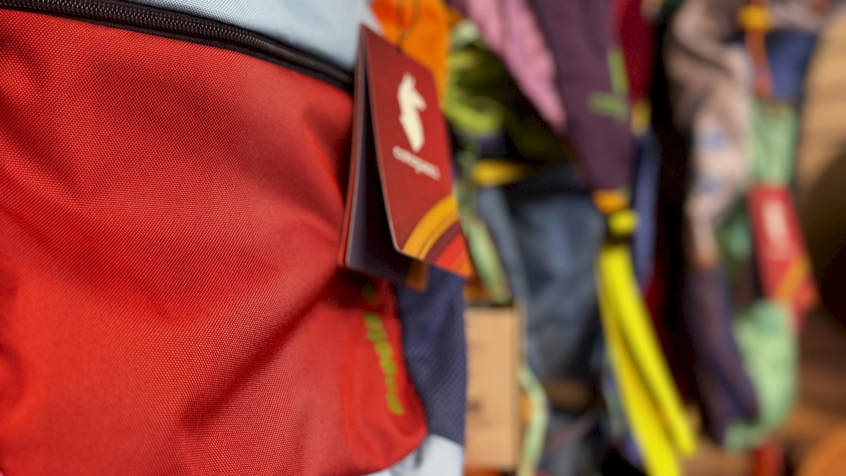 The image shows a display of colorful backpacks hanging in a store, with some visible tags and brand logos on the bags.